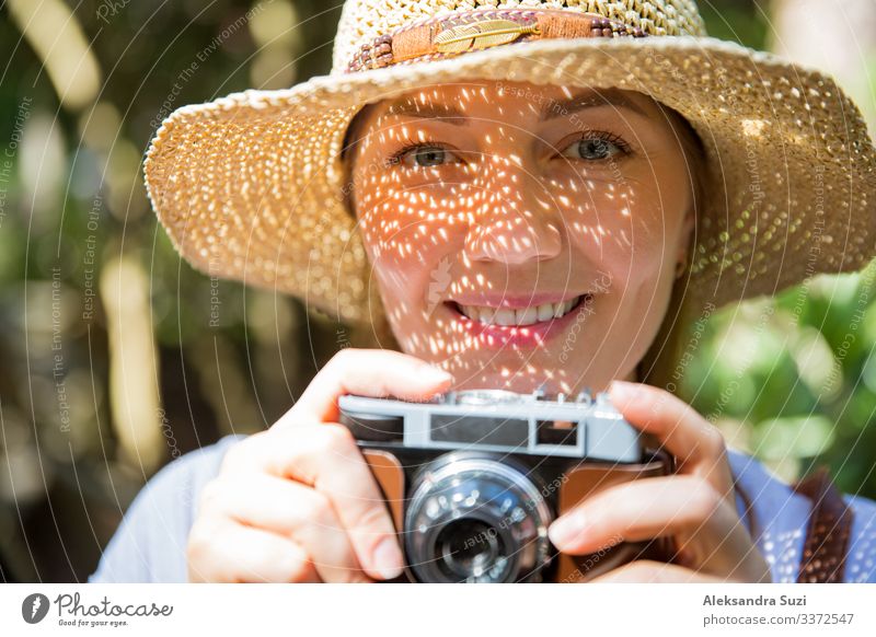 Nahaufnahme einer schönen Frau mit Strohhut, die im Tropenwald reist und mit einer Retro-Kamera fotografiert. Leichte Schatten durch die ausgeschnittene, detaillierte Krempe im Gesicht. Tourist mit Rucksack.