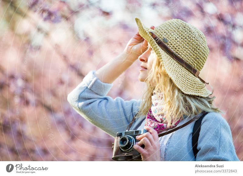 Porträt einer schönen Frau mit Strohhut, die in einem schönen Park mit blühenden Kirschbäumen reist und Fotos mit einer Retro-Kamera macht. Tourist mit Rucksack.