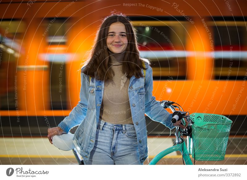 Teenager in Jeans mit gelbem Rucksack und Fahrrad in der U-Bahn-Station stehend, auf den Zug wartend, lächelnd, lachend. Oranger Zug fährt hinter dem Mädchen vorbei. Futuristischer U-Bahnhof. Finnland, Espoo