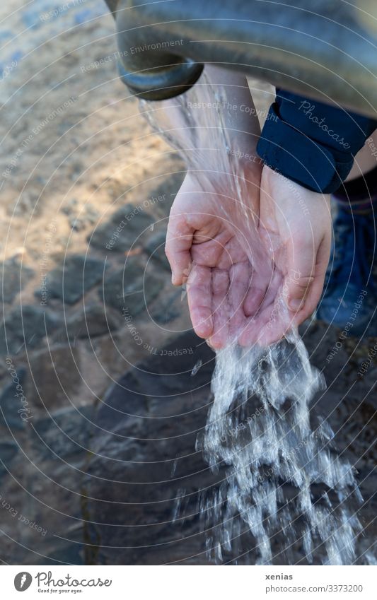 Hände im Strahl der Wasserpumpe mit Blick auf unscharfem Kopfsteinpflaster Trinkwasser Hand 1 Mensch Wasserstrahl frisch nass grau Gedeckte Farben Außenaufnahme