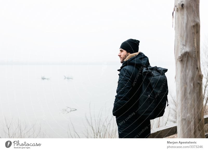 Junger Mann steht am Wintermorgen am Fluss aktiv Aktivität Erwachsener Abenteuer allein Rucksack lässig Küste kalt Landschaft Tag genießen Entdecker neblig