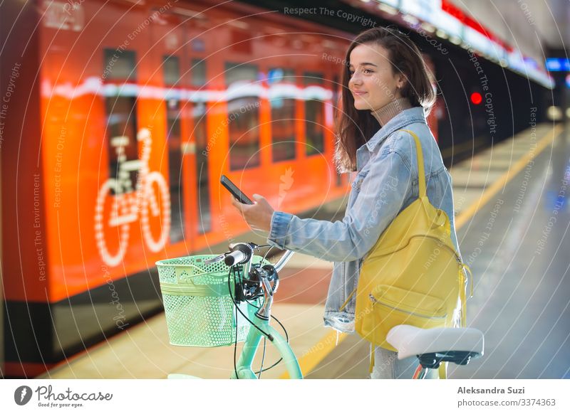 Teenager mit Rucksack und Fahrrad auf der U-Bahn-Station stehend, Smartphone in der Hand haltend, blätternd und simsend, lächelnd und lachend. Futuristisch helle U-Bahn-Station. Finnland, Espoo