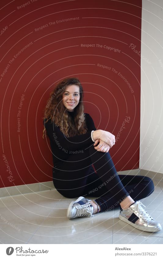 Junge Frau sitzt an einer roten Wand Lifestyle Stil Freude schön Leben Wohlgefühl Wohnung Jugendliche 18-30 Jahre Erwachsene Jeanshose Pullover Turnschuh