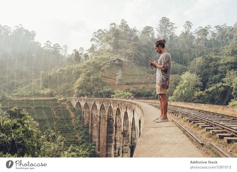 Mann im Urlaub surft mit Smartphone auf der Brücke Tourismus reisen Browsen zuschauend fotografieren Landschaft tropisch benutzend Handy Sri Lanka Asien grün