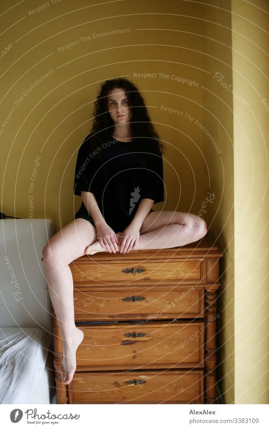 Junge Frau sitzt auf einer Kommode vor einer gelben Wand Blick in die Kamera Porträt Zentralperspektive Schwache Tiefenschärfe Tag Textfreiraum rechts