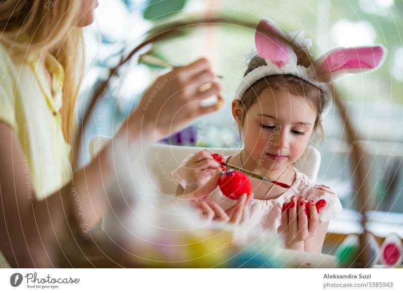 Eine Mutter und eine Tochter feiern Ostern und bemalen Eier mit dem Pinsel. Eine glückliche Familie, die lächelt und lacht. Ein süßes kleines Mädchen in Hasenohren, das den Feiertag vorbereitet.