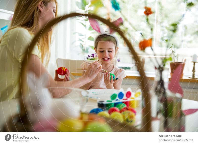 Eine Mutter und eine Tochter feiern Ostern und bemalen Eier mit dem Pinsel. Eine glückliche Familie, die lächelt und lacht. Ein süßes kleines Mädchen in Hasenohren, das den Feiertag vorbereitet.