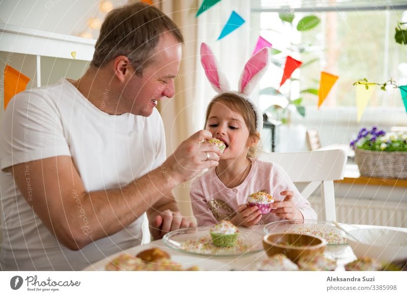Vater und Tochter feiern Ostern und essen mit Glasur überzogene Törtchen. Fröhlicher Familienurlaub. Süßes kleines Mädchen in Hasenohren. Wunderschön dekorierter Raum