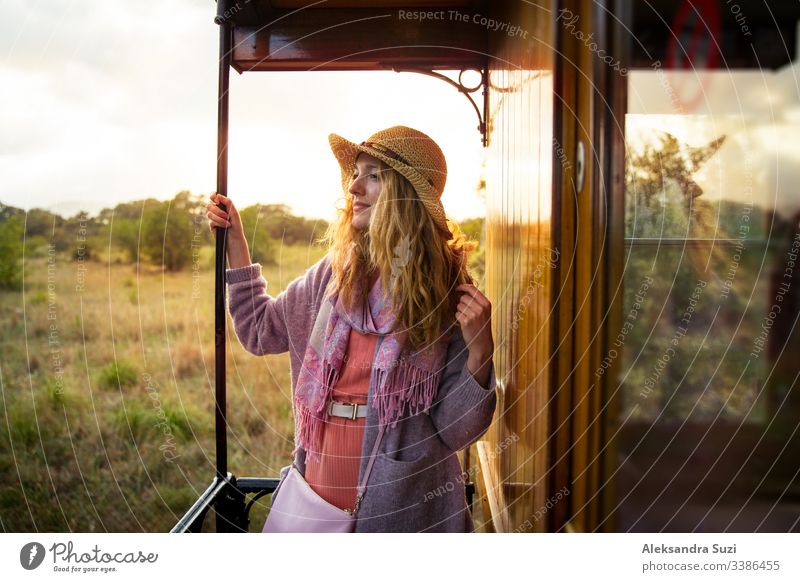 Junge Frau mit Strohhut reist mit einem Retro-Holzzug. Sonnenuntergangslandschaft, Wind in blonden Haaren. Mädchen lächelt fröhlich. Mallorca, Spanien.