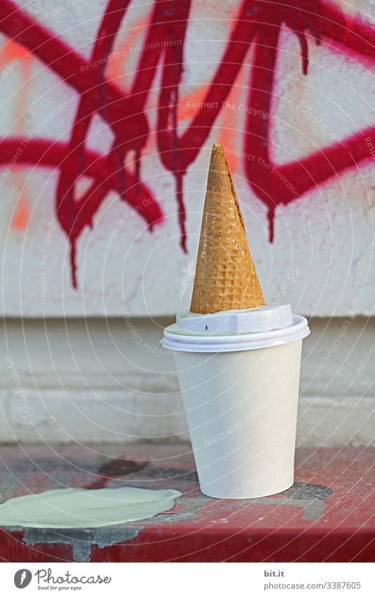 Eis hinterläßt Klecks und Sauerei. Leere Eiswaffel liegt nachhaltig, auf dem Kopf auf einem leeren, weißem Kaffeebecher von einem Coffee to go. Bunte Graffiti verziert die Wand im Hintergrund.