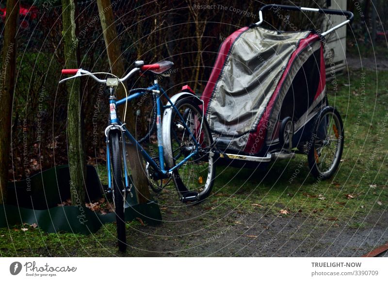 Blau-silbernes Fahrrad mit roten Handgriffen und Anhänger (Buggy) in grau / magenta für Kindertransport bei Regen am Gartenzaun abgestellt Fahrradanhänger