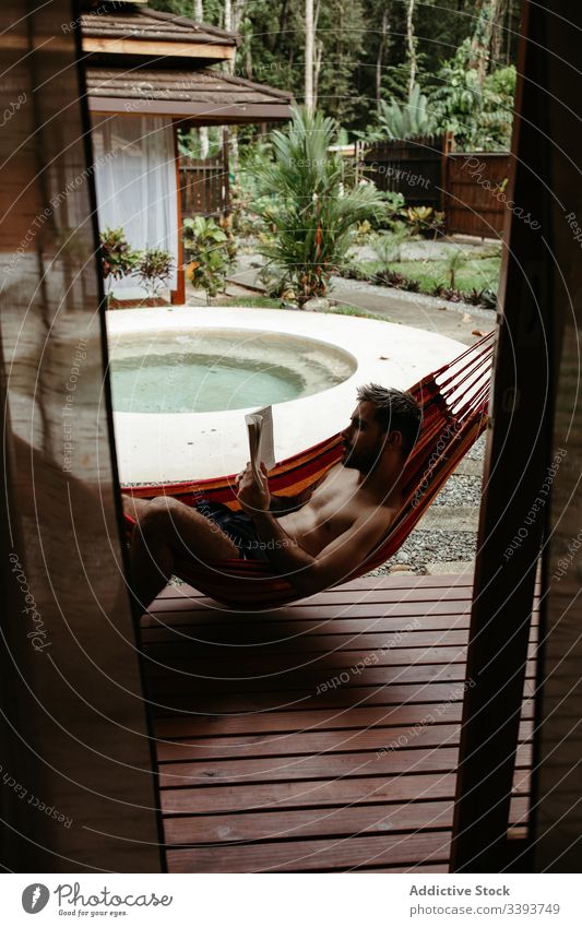 Junger männlicher Tourist in Bademode mit Buch auf Hängematte Mann Resort Hotel räkeln Kälte Beckenrand lesen exotisch tropisch Badeanzug sich[Akk] entspannen
