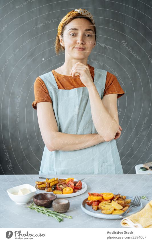 Zuversichtliche Frau steht am Tisch mit gebratenem Gemüse Koch Küche Speise Mittagessen Teller Lebensmittel Vorbereitung heimwärts Mahlzeit Bestandteil gebacken