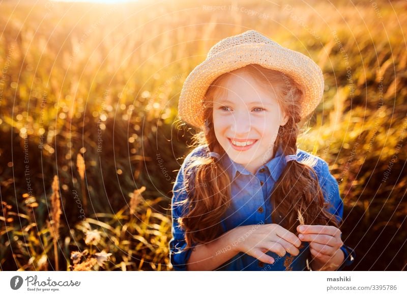 glückliches Mädchen in blauem Kleid und Stroh, das auf einer sonnigen Sommerwiese spazieren geht. Lifestyle-Aufnahme, ländliches Leben und Sommerreisekonzept