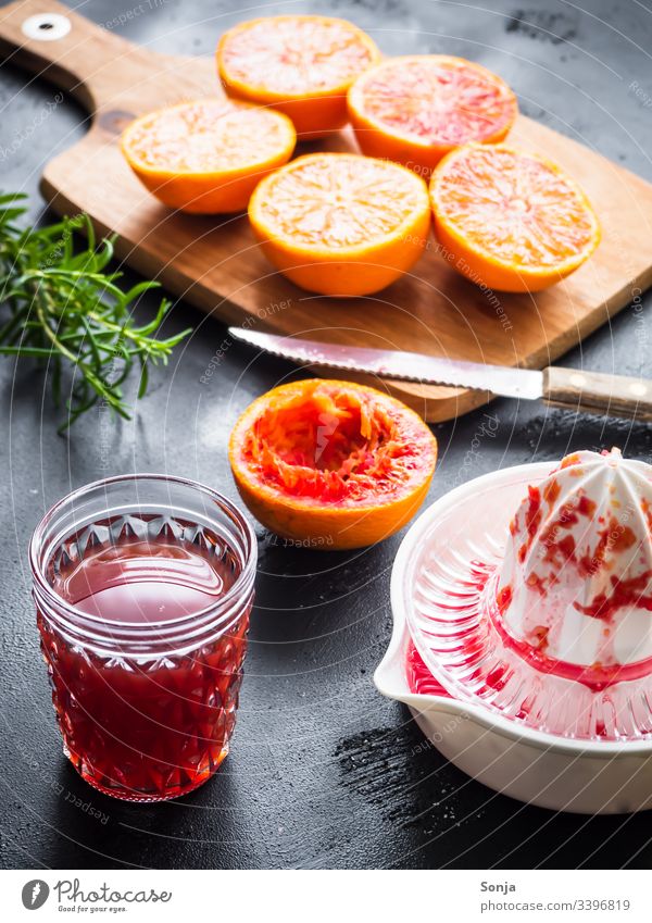 Frisch gepresster Blutorangensaft in einem Glas, eine Saftpresse und ein Schneidebrett mit halbierten Orangen auf einem rustikalen Hintergrund blutoragensaft