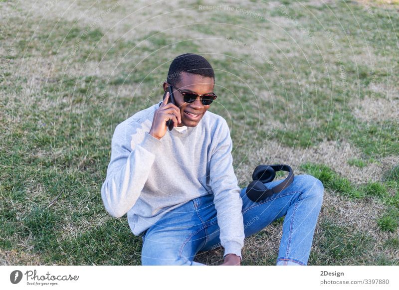 Modischer schwarzer Mann mit Sonnenbrille, der im Freien sitzt und seinen Nachrichten-Feed oder seine Nachrichten über soziale Netzwerke per Handy abruft