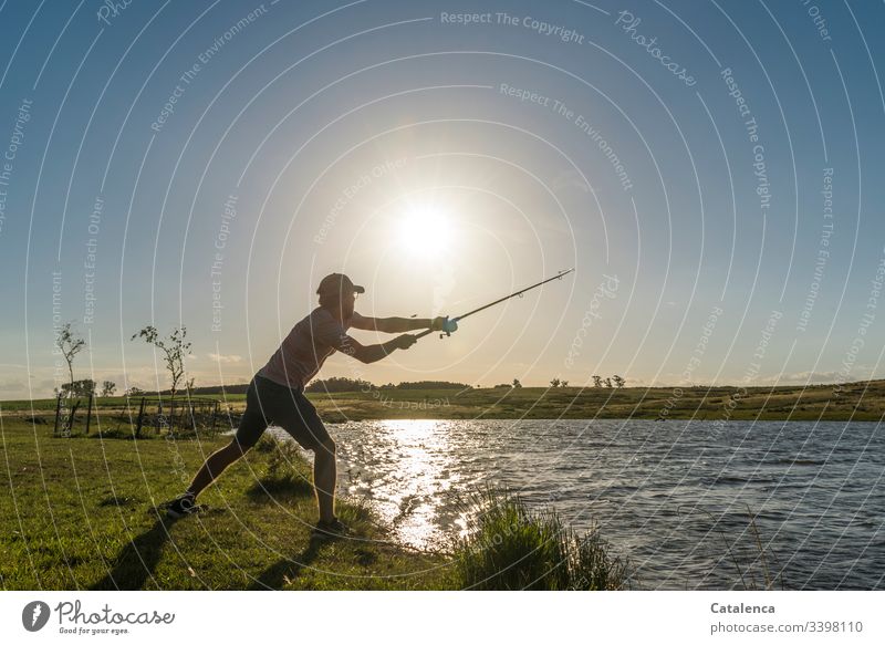 Junger Mann wirft am Seeufer eine Angel aus, die Sonne scheint, das Wasser glänzt Angeln Fisch Fischer Angelrute Farbfoto Küste Lebensmittel Angler