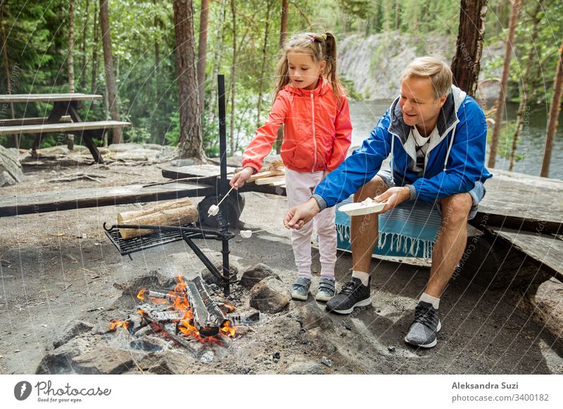 Mann und seine kleine Tochter grillen im Wald am felsigen Seeufer, machen ein Feuer, grillen Brot, Gemüse und Marshmallow. Familie erkundet Finnland. Skandinavische Sommerlandschaft.