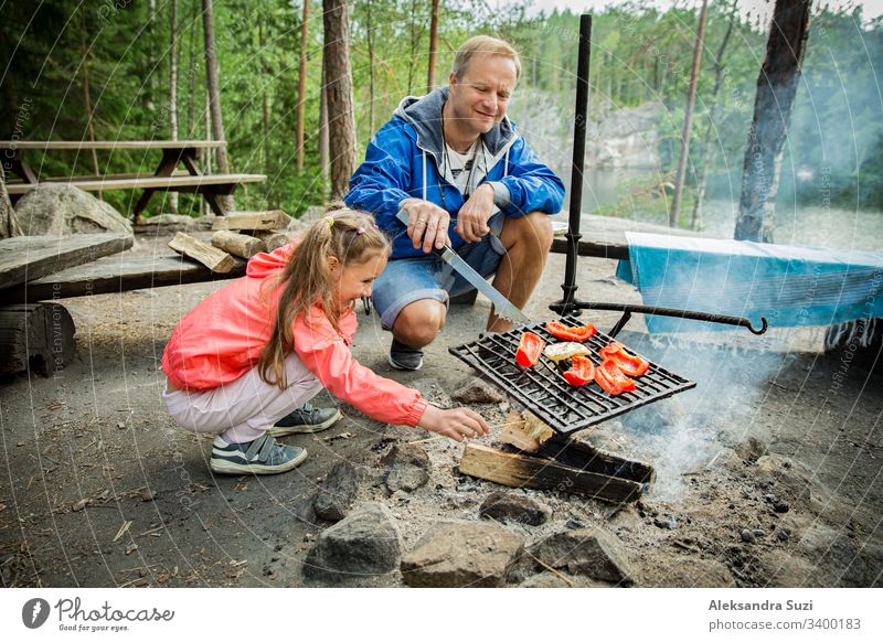 Mann und seine kleine Tochter grillen im Wald am felsigen Seeufer, machen ein Feuer, grillen Brot, Gemüse und Marshmallow. Familie erkundet Finnland. Skandinavische Sommerlandschaft.