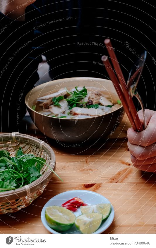 Eine Schüssel vietnamesische Pho Suppe mit frischen Kräutern und einer Person mit Stäbchen in der Hand beim Essen Pho Bo Vietnam gesund Urlaub heiß limetten