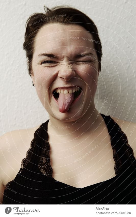 Portrait einer jungen Frau - witziger Ausdruck mit Zunge rausstrecken einzigartig Blick in die Kamera positiv Glück natürlich Farbfoto braune Augen kinngrübchen
