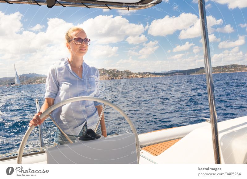 Attraktive blonde Skipperin, die das schicke Katamaran-Segelboot an einem sonnigen Sommertag auf ruhigem blauen Meerwasser steuert. nautisch Lenkrad Boot Frau