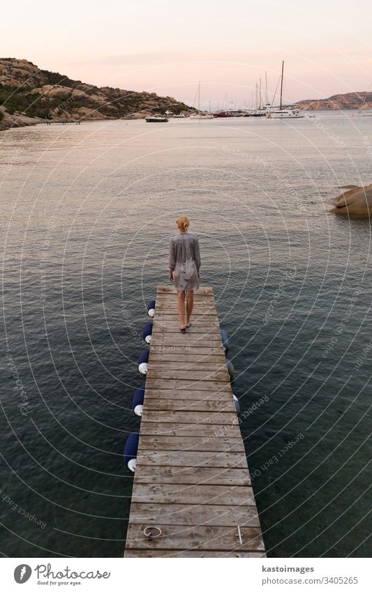 Schöne Frau in luxuriösem Sommerkleid, die auf einem Holzsteg steht und in der Abenddämmerung die friedliche Meereslandschaft genießt. Reisende Frau steht auf einem hölzernen Pier in Porto Rafael, Costa Smeralda, Sardinien, Italien.