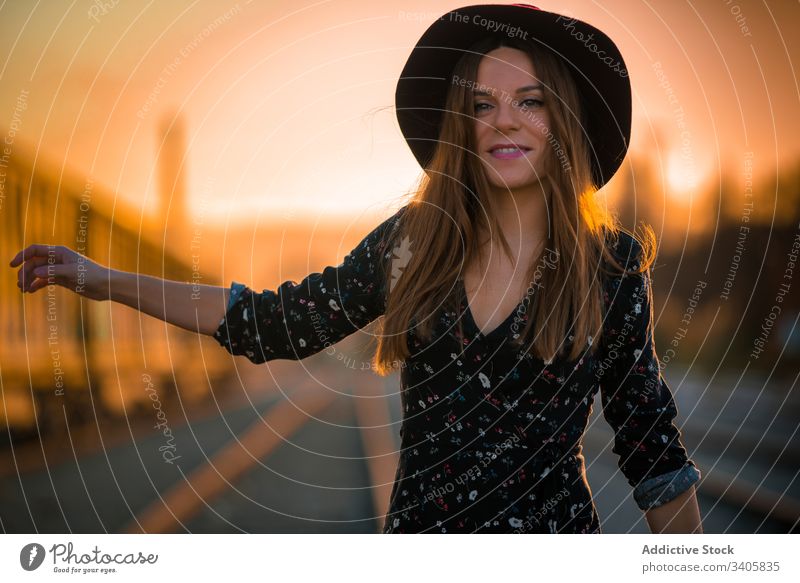 Fröhliche Frau trampt bei Sonnenuntergang auf der Eisenbahn trampen Straße Landschaft Lächeln gestikulieren reisen Stil Hut Lifestyle Ausflug Route Weg Autobahn