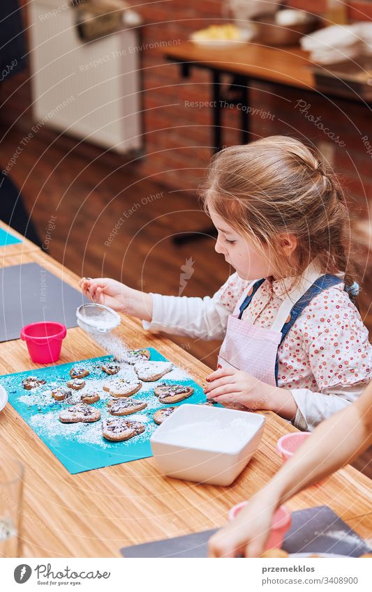 Kleines Mädchen verziert ihre gebackenen Kekse mit bunten Streuseln und Puderzucker. Kind, das am Backworkshop teilnimmt. Backkurse für Kinder, aufstrebende kleine Köche. Kochen lernen. Kombinieren und Rühren von vorbereiteten Zutaten.