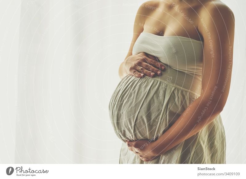Junge schöne schwangere Frau steht zu Hause am Fenster Schwangerschaft Baby Bauch heimwärts Mutterschaft erwartend Glück Licht Kleid Konzept Pflege Person Leben