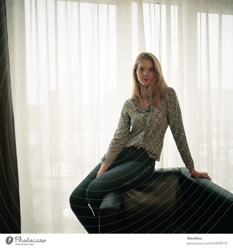 Portrait einer jungen Frau auf einem Sessel vor dem Fenster Mädchen blond schön schlank anmutig elegant lifestyle Wohnen Wohnung zu Hause Gardine Bluse Muster