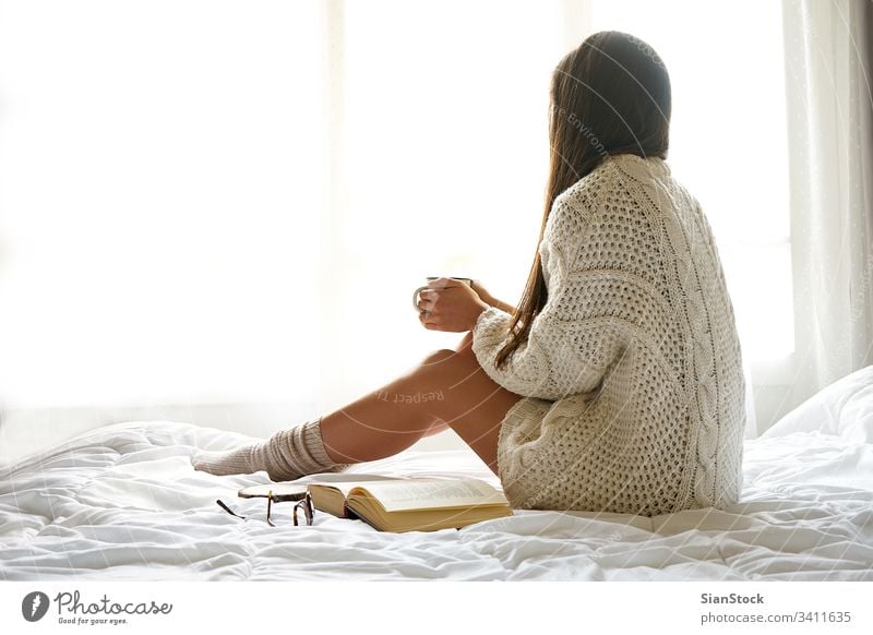 Weiches Foto einer Frau auf dem Bett mit einem alten Buch und einer Tasse Kaffee lesen Fenster Ansicht LAZY Sonntag Winter Tee Morgen Mädchen heimwärts