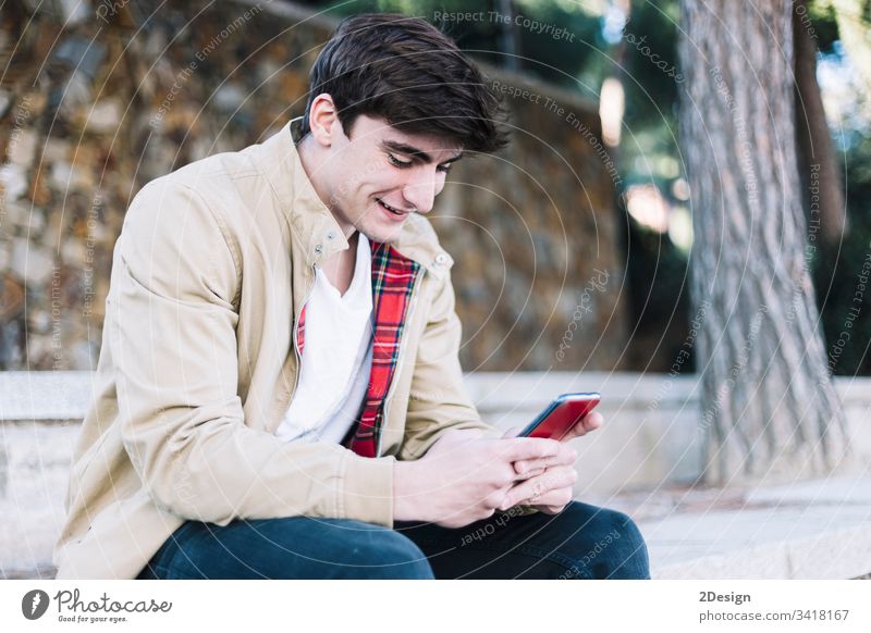 Junger hispanischer Mann, der im Freien sitzend ein Mobiltelefon benutzt männlich Sitzen jung 1 Telefon Mobile Beteiligung Menschen photogen modern Typ Person