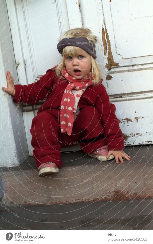 Kleines, blondes, sprechendes  Mädchen in Winterkleidung, klettert auf einer Treppe, vor einer alten, weißen Haustür aus Holz. Kind klein Kleinkind Spielen