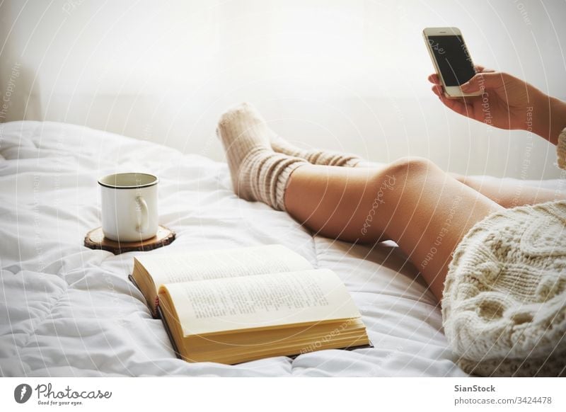 Weiches Foto einer Frau auf dem Bett mit einem alten Buch, einer Tasse Kaffee und einem Smartphone lesen Fenster Ansicht LAZY Sonntag Winter Tee Morgen Mädchen