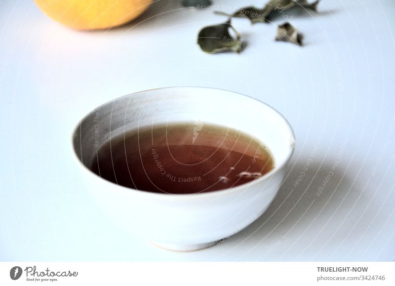 Frisch aufgebrühter Asssam Tee in heller chinesischer Keramik Schale auf weißem Tisch mit einigen herab gefallenen Blättern und einer Grapefruit- Schwarzer Tee
