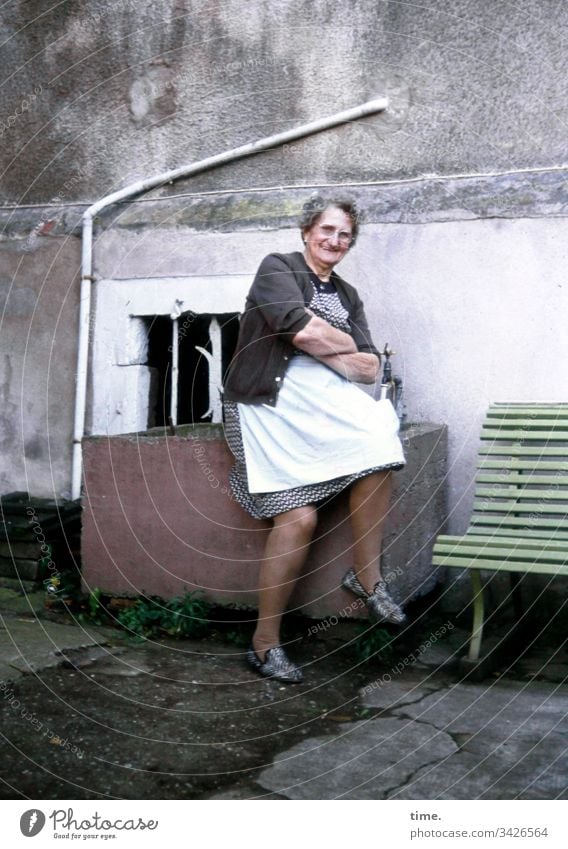 Kraft & Güte Frau Seniorin Hinterhof Schürze sitzen Bank Abflussrohr Beton Wand Mauer wohnen Fenster freundlich Strickjacke lächeln Kleid verschränkte Arme