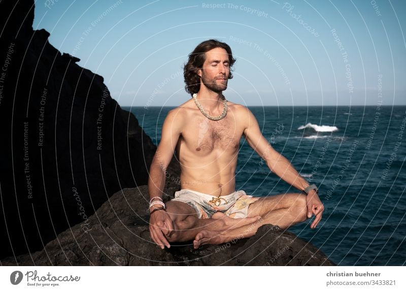 junger mann am meer in yoga position nackt lange haare 35 jahre fels badehose hippie blauer himmel sport wellness relax urlaub frieden ruhe pilates