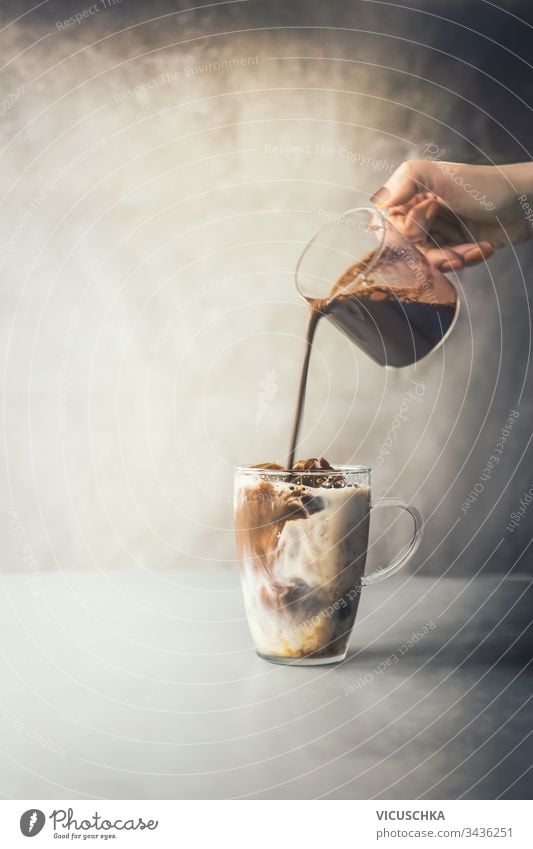 Barista gießt von Hand Kaffeesahne in einen Glasbecher mit geeistem Kaffeelatte auf einen rustikalen Tisch vor einem Betonwandhintergrund. Zubereitung von Eiskaffee. Erfrischendes Sommergetränk. Kaltes Getränk. Zubereitung von Eiskaffee.