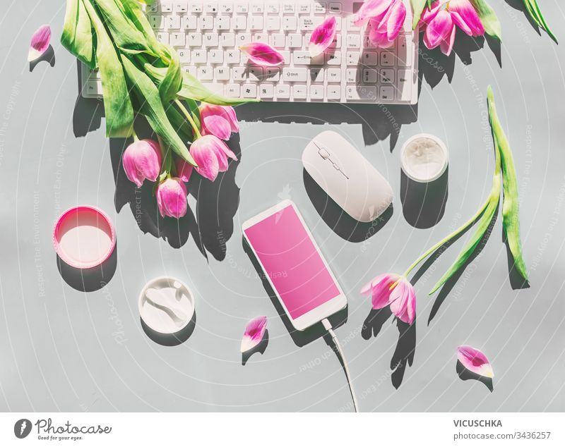 Weiblicher Schreibtisch mit hellrosa Tulpenblumenstrauß, Kosmetik, PC-Tastatur, Smartphone mit leerem Bildschirm und Kabel . Ansicht von oben. Muttertag . Frühling . Begrüßung