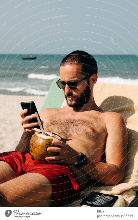 Gestrandeter Mann im Urlaub sitzt am Strand mit einer Kokosnuss in der einen Hand und ist mit seinem Smartphone beschäftigt Urlauber gestrandet Reisen Tourist