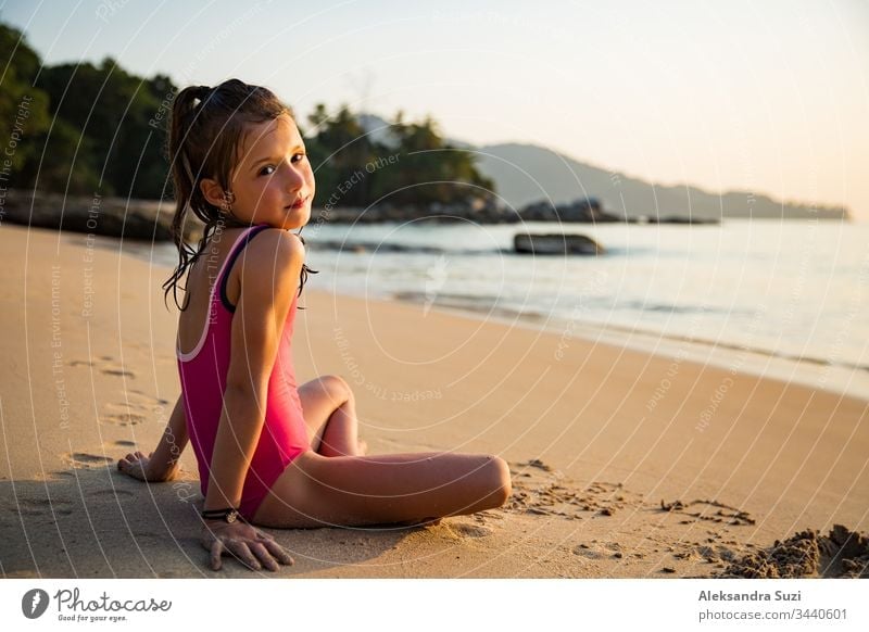 Süßes fröhliches kleines Mädchen, das im Badeanzug am Strand mit Sand spielt, ein Herz zeichnet und schreibt. Wunderschöner Sommersonnenuntergang, Meer, Kokospalmen, malerische exotische Landschaft. Phuket, Thailand