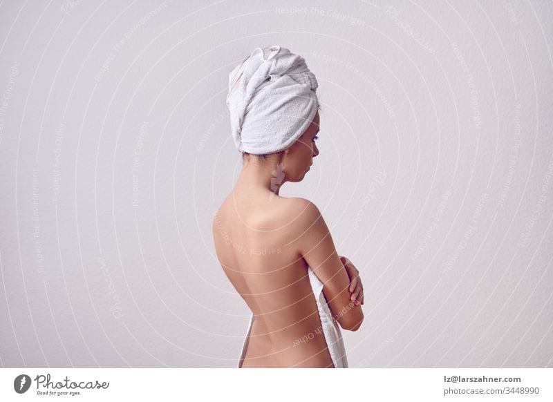 Dünne junge Frau nach dem Duschen von ihrem Rücken mit einem weißen Handtuch über dem Kopf und mit nacktem Rücken. Halbfigur vor schlichtem hellgrauen Hintergrund