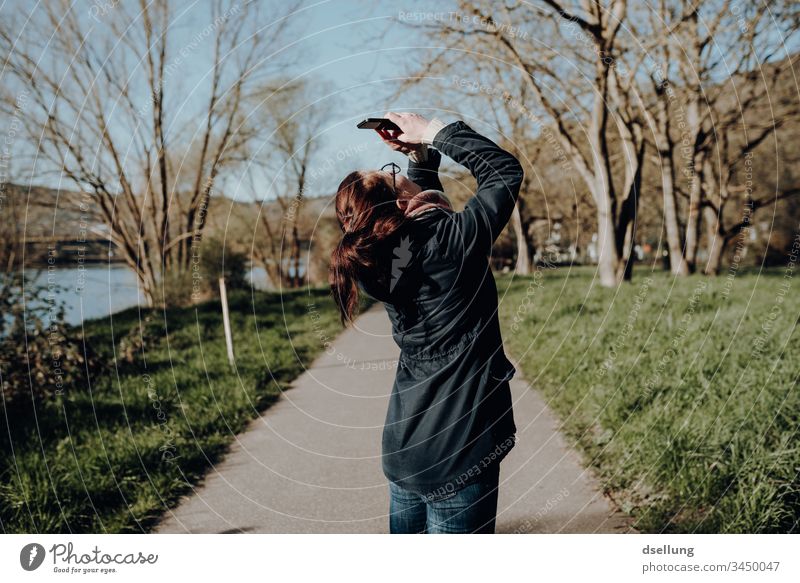 Junge Frau in der Natur macht Fotos mit dem Smartphone, wobei sie ihren Rücken in einer Position hält, die Jedem Orthopäden Schweißflecken unter die Achseln zaubert