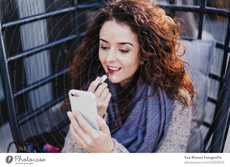 Porträt einer jungen schönen Frau, die draußen auf einer Terrasse roten Lippenstift benutzt. Sie benutzt ein Mobiltelefon als Spiegel. Make-up und Lifestyle-Konzept