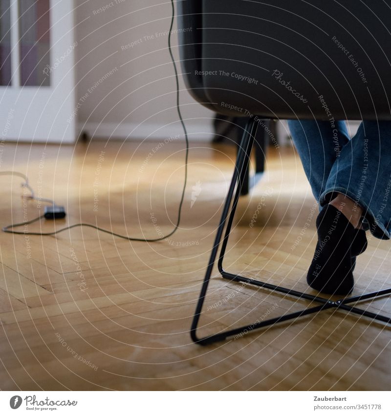 Beine einer Frau, die auf einem Stuhl im Homeoffice sitzt, mit Kabel und Netzteil ihres Computers, auf Parkett sitzen Füße Socken Netzkabel zuhause Strümpfe
