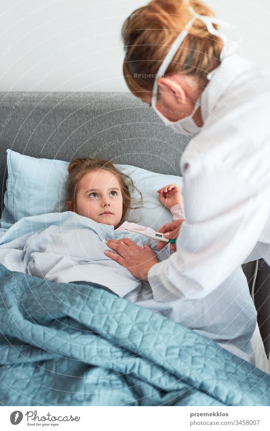 Der Arzt besucht den kleinen Patienten zu Hause. Messung der Temperatur eines kranken Mädchens, das im Bett liegt. Frau trägt Uniform und Gesichtsmaske. Medizinische Behandlung