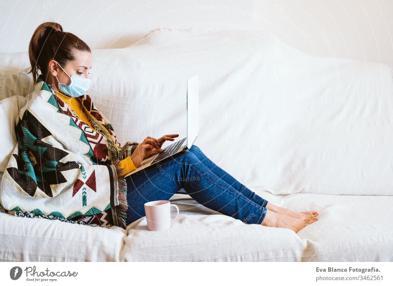 junge Frau, die zu Hause am Laptop arbeitet, auf der Couch sitzt und eine Schutzmaske trägt. Das Konzept "zu Hause bleiben" während des Coronavirus covid-2019