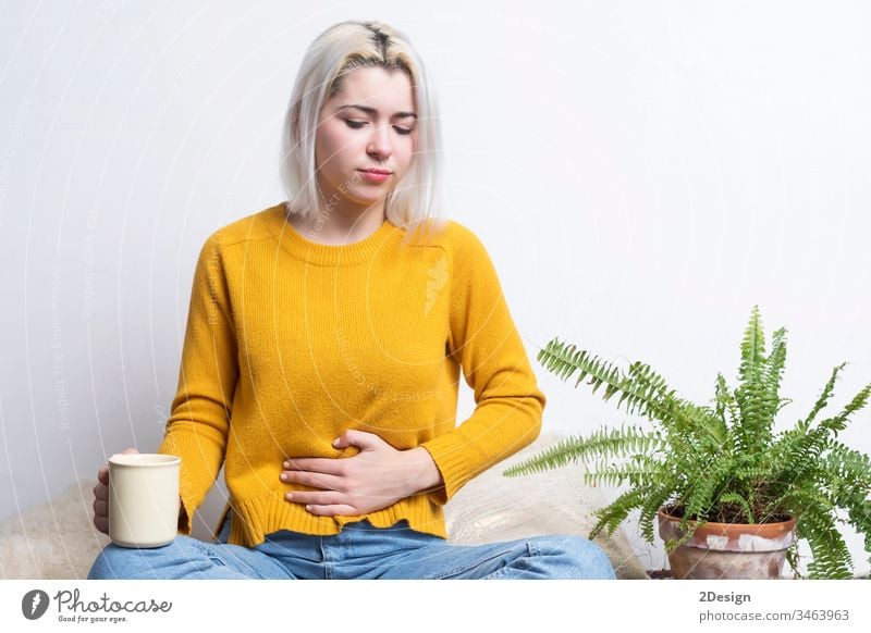 Junge blonde Frau sitzt zu Hause und hält eine Tasse Tee, während sie ihre Hand in den Bauch steckt Gesundheitswesen ungesund Menses Mediziner Sitzen krank