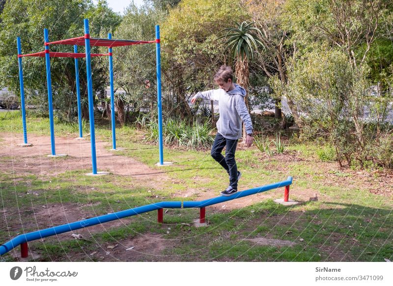 Junge (Kind), der auf einem Spielplatz auf einer niedrigen Stange balanciert Ausgewogenheit sonnig im Freien Sträucher Gras